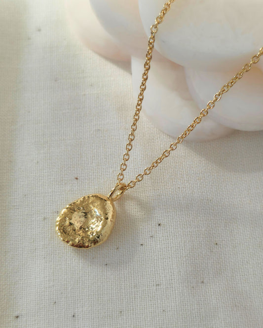 A gold chain necklace with a textured oval-shaped gold pendant, displayed on a light beige linen fabric background