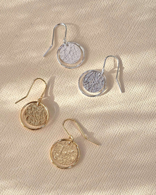 Two pairs of handmade gold and silver drop earrings with textured circles framed by delicate wire loops lying on paper