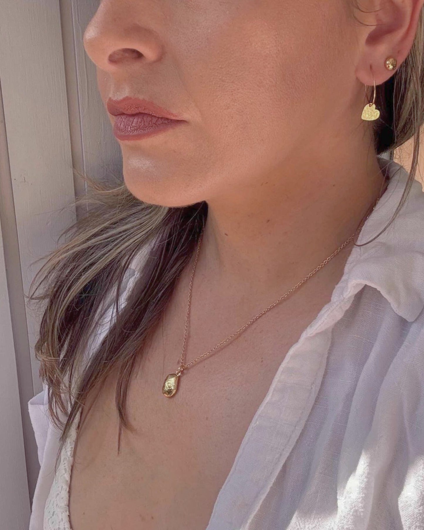 Close up of a woman wearing a gold necklace with an oval pendant and gold heart shaped earrings