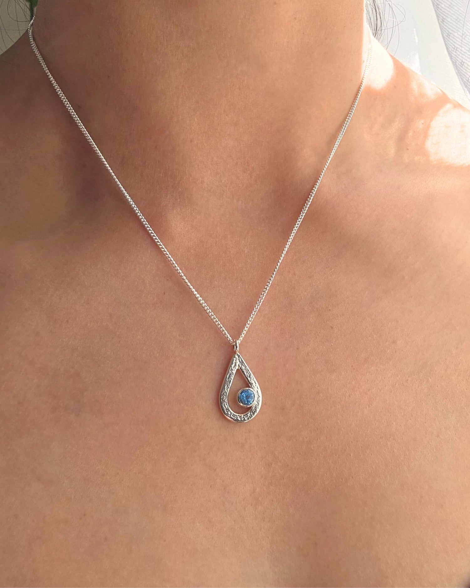 Silver teardrop necklace with a textured surface set with a blue topaz gemstone on a person's neck