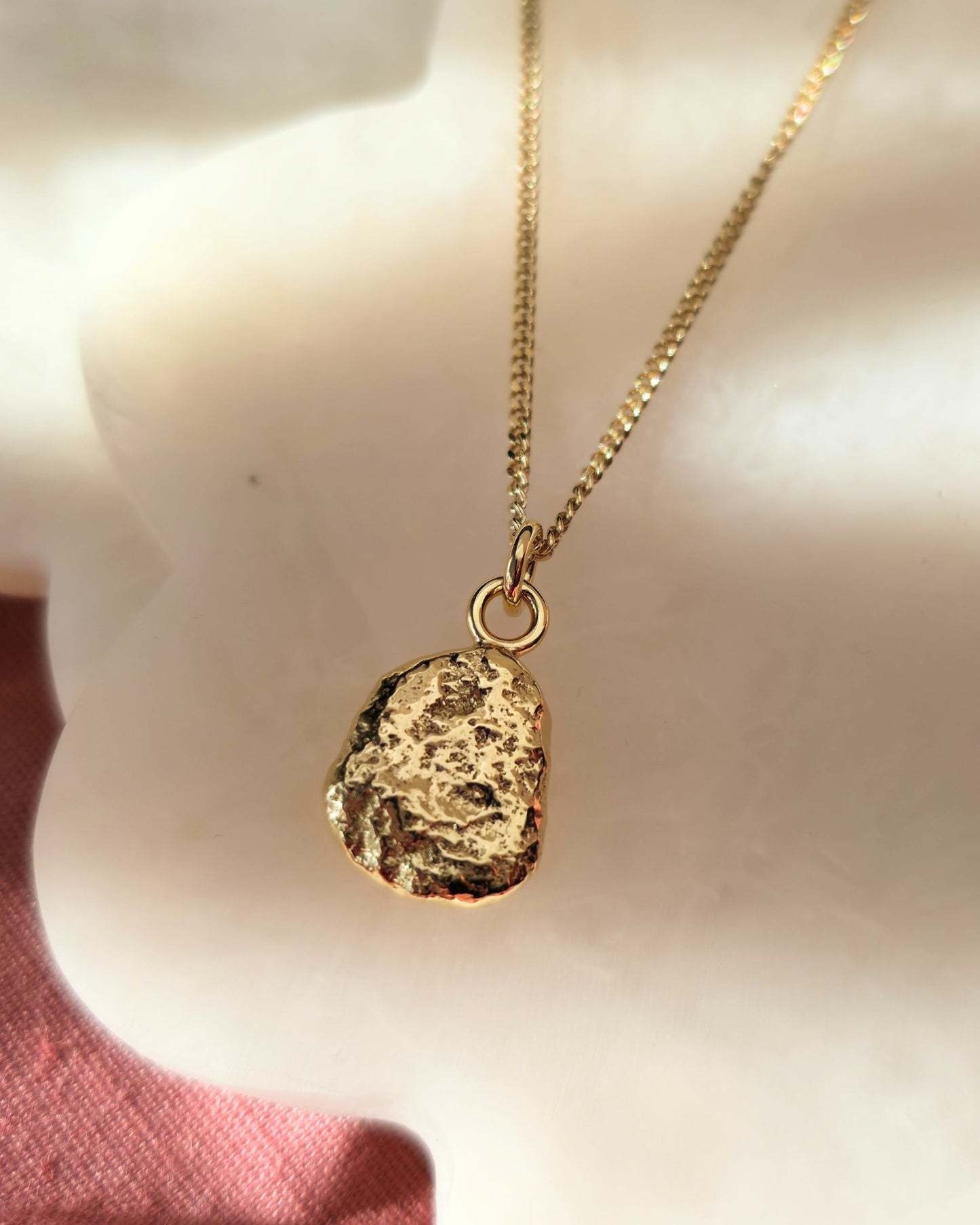 Gold necklace with a textured pendant lying in diffused sunlight on a cream surface.