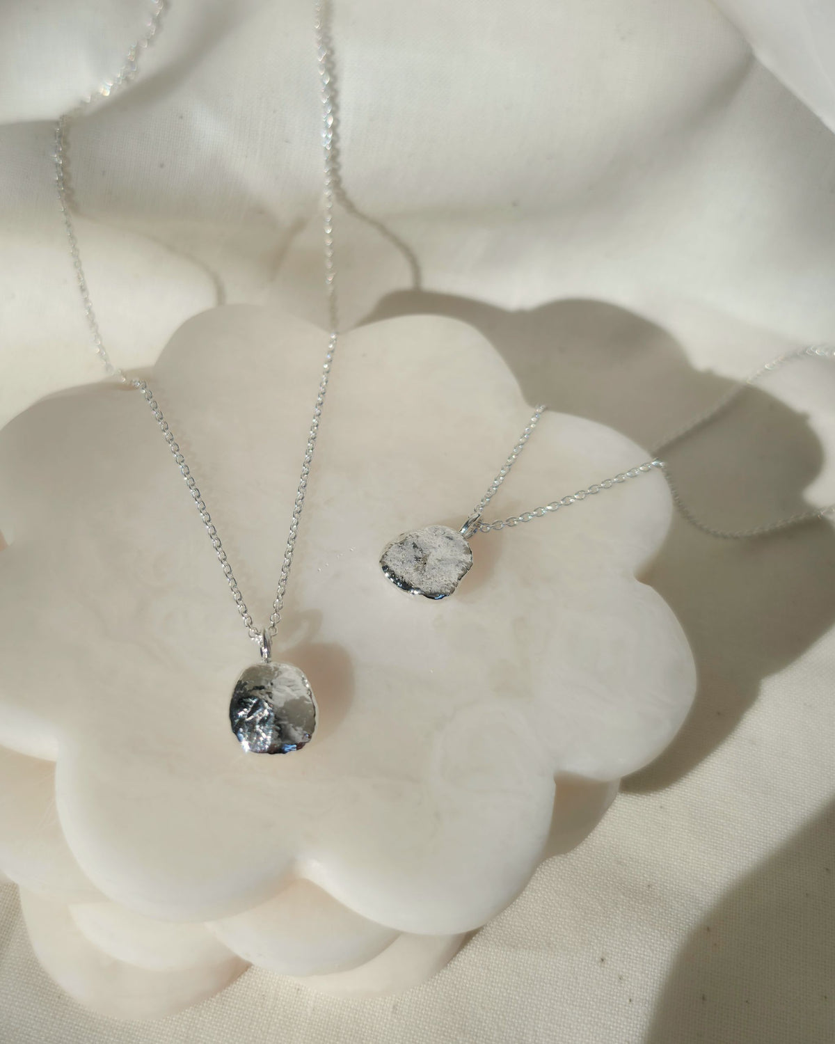 Two silver chain necklaces with oval-shaped textured pendants, displayed on a white flower-shaped stand against a soft white fabric background