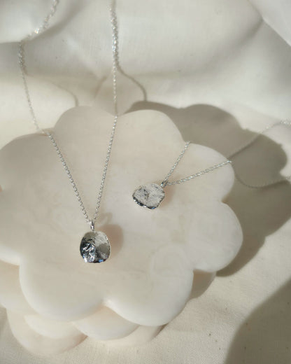 Two silver chain necklaces with oval-shaped textured pendants, displayed on a white flower-shaped stand against a soft white fabric background