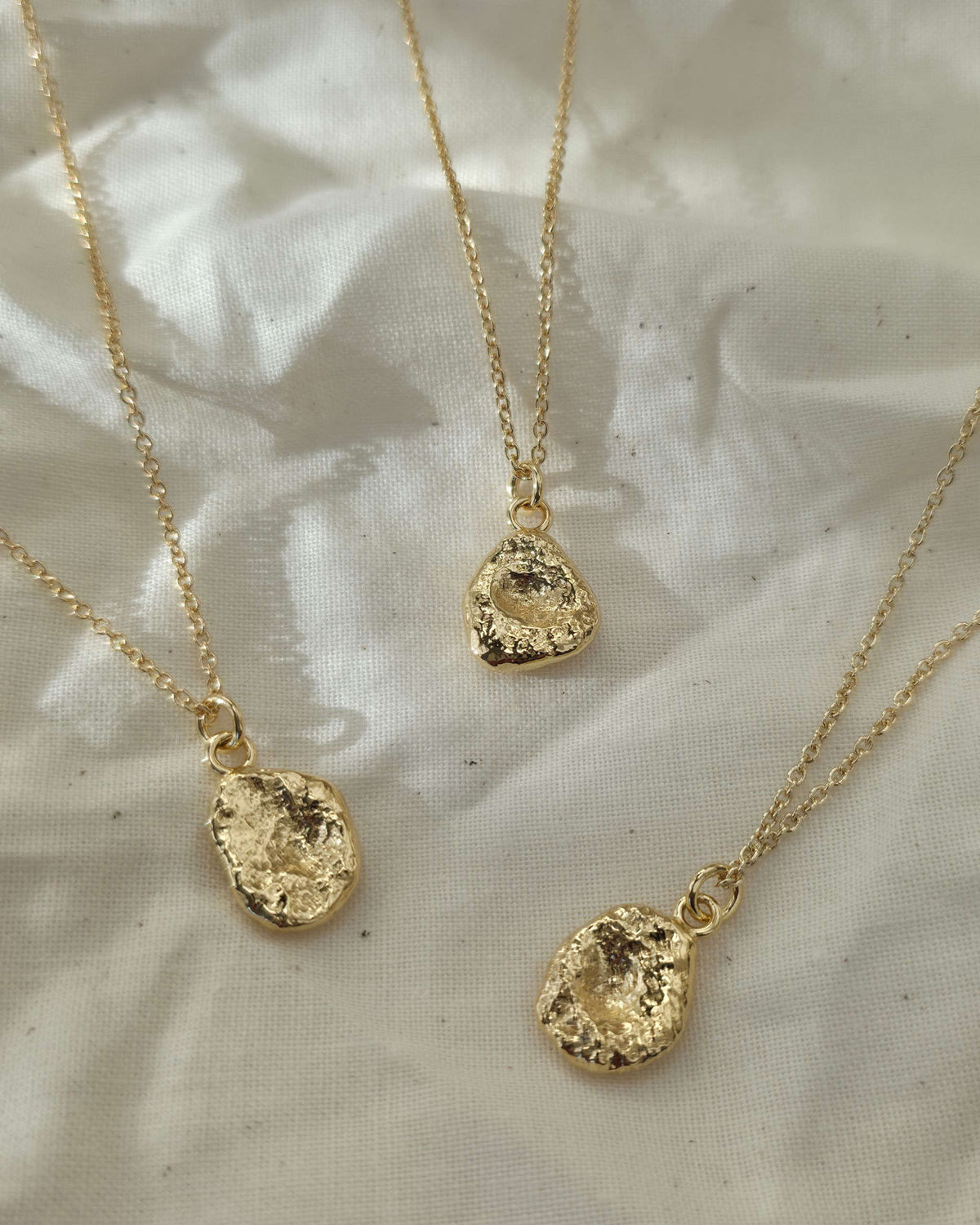 Three gold necklaces with textured pendants lying on cream coloured fabric