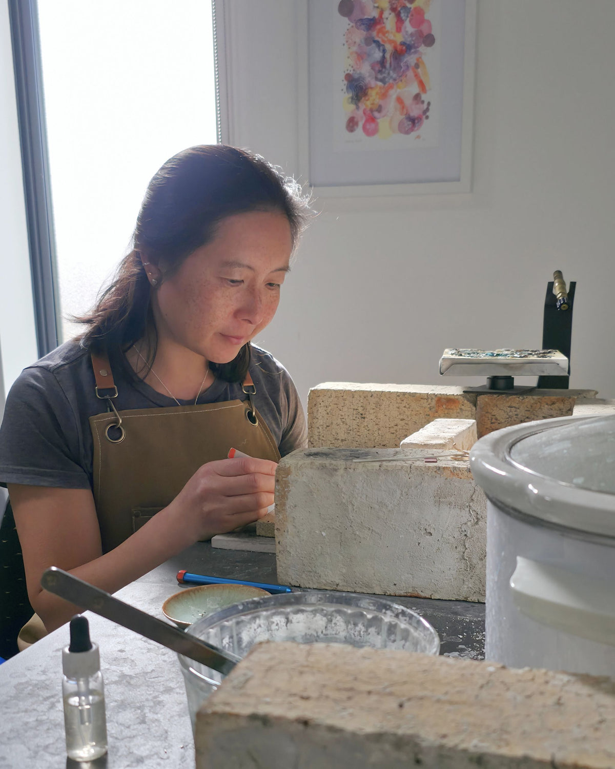 A woman wearing a brown apron over a grey t-shirt sits at a jewellery bench with tweezers in her hand. In front of her are several textured, light-coloured stone blocks, a small spray bottle, and a white ceramic pot.