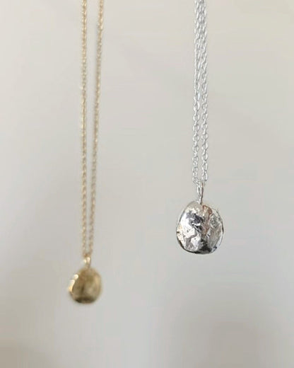 Two textured round pendant necklaces in gold and silver, hanging against a light background