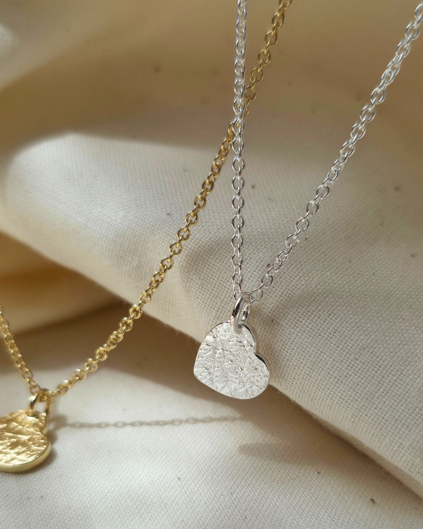 Two heart-shaped pendants on gold and silver chains against a beige fabric background