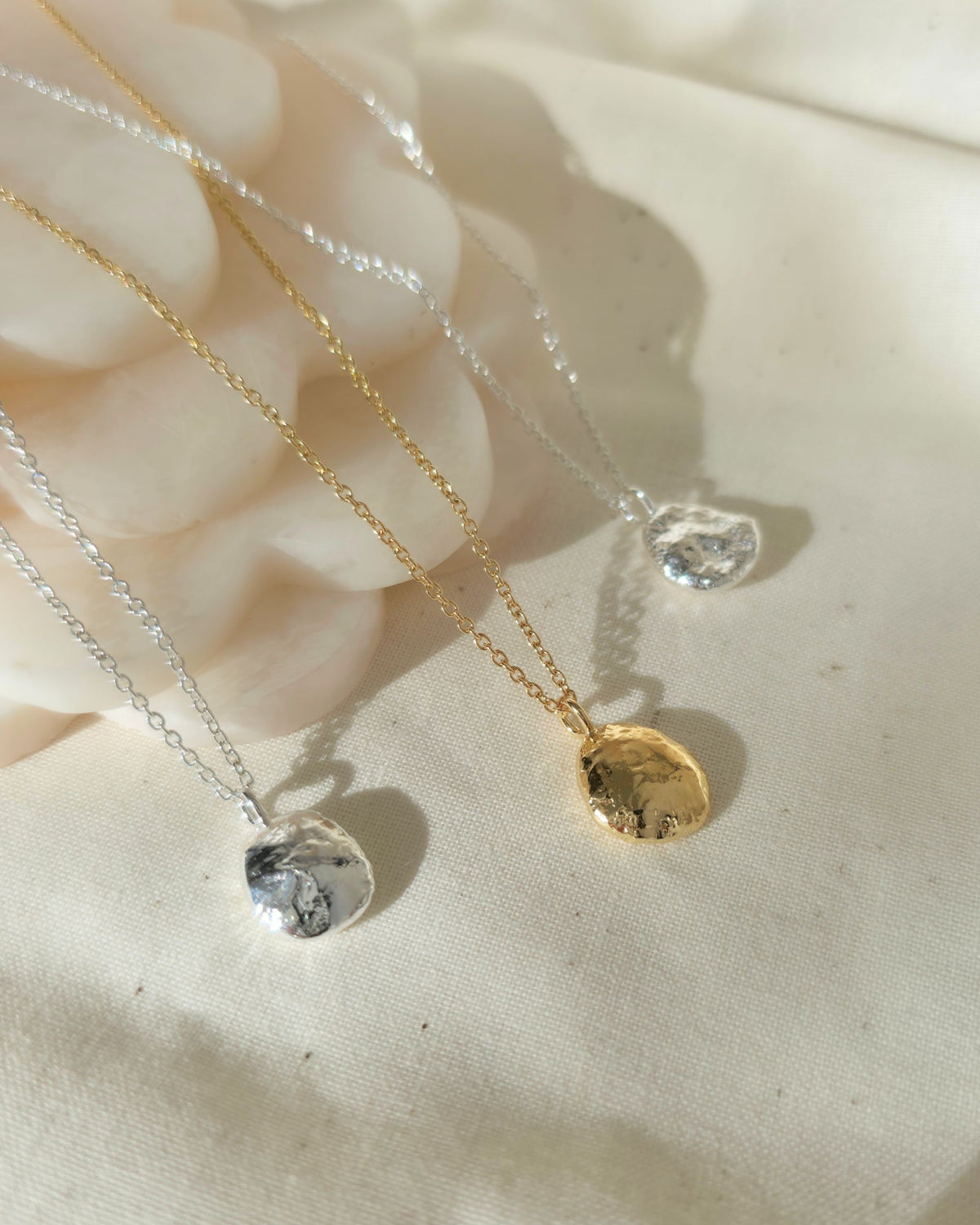 Three delicate pendant necklaces with textured round pendants in gold and silver tones, displayed on a soft cream fabric background