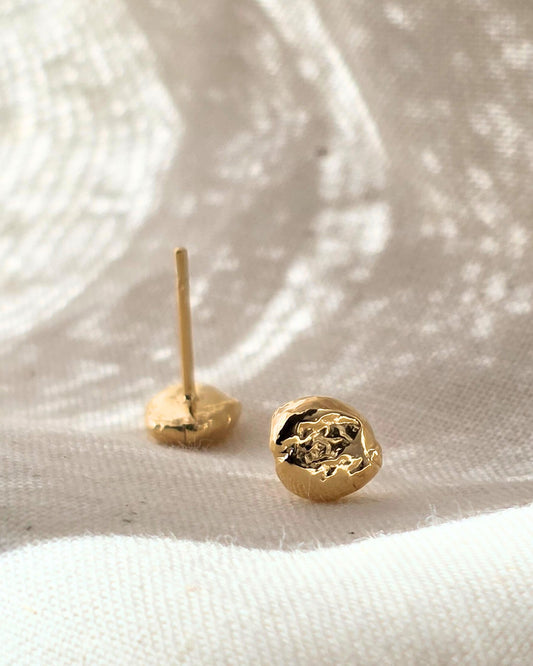 Organic molten gold stud earrings with a rippled texture lying in shadow on beige fabric background