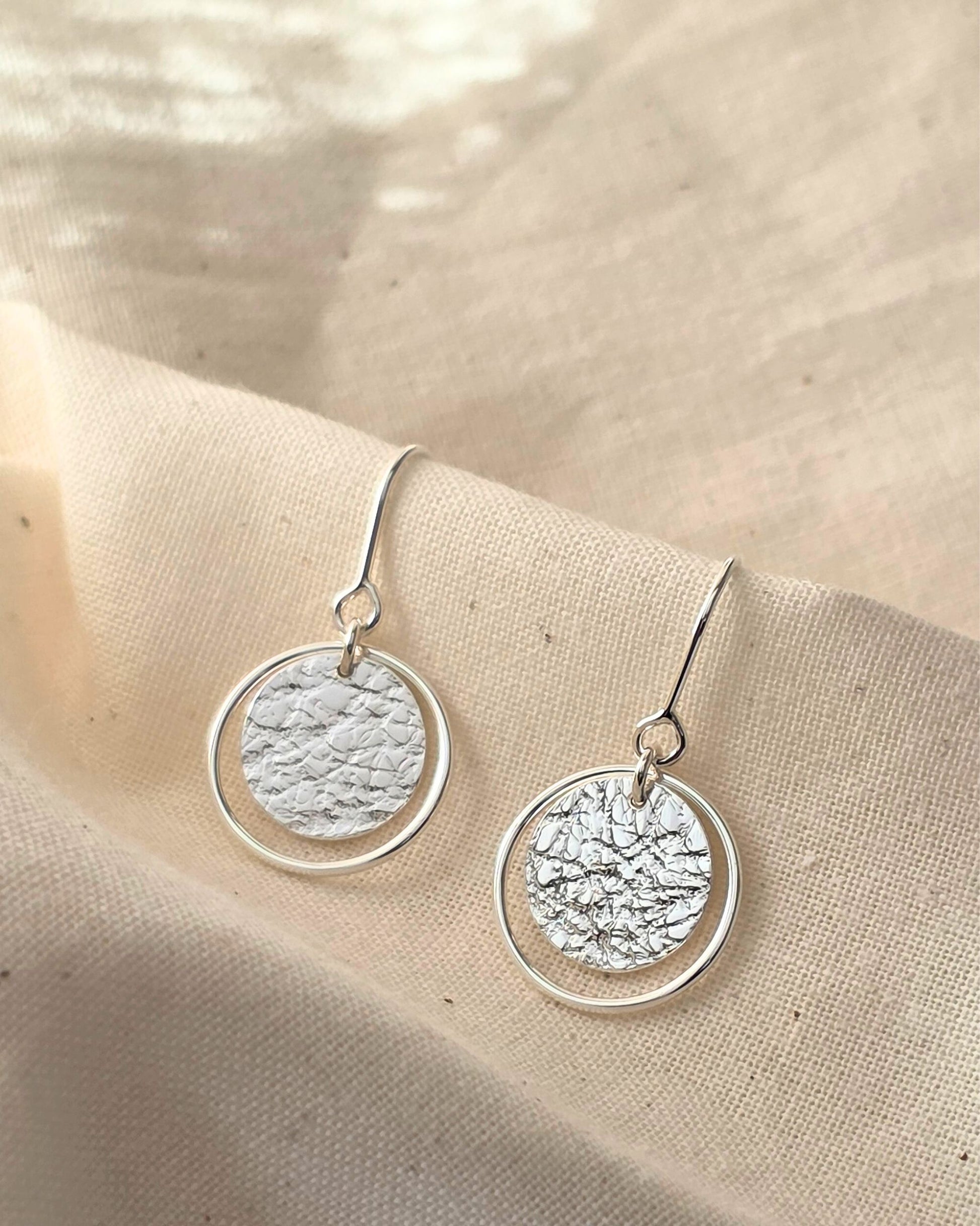 Handmade earrings with textured circles framed within thin sterling silver loops lying on beige fabric