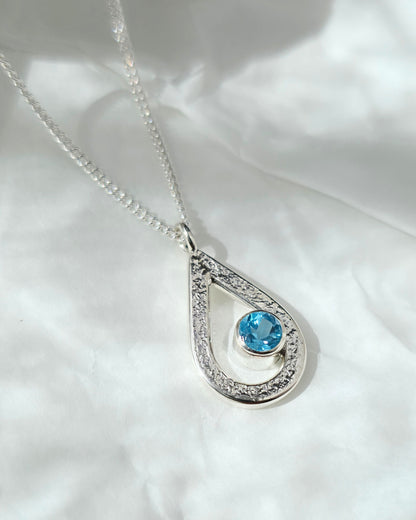 Textured silver teardrop pendant with a blue topaz gemstone on a white fabric background