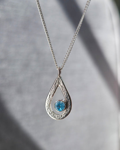 Teardrop-shaped pendant with a blue  topaz gemstone on a chain against a grey background
