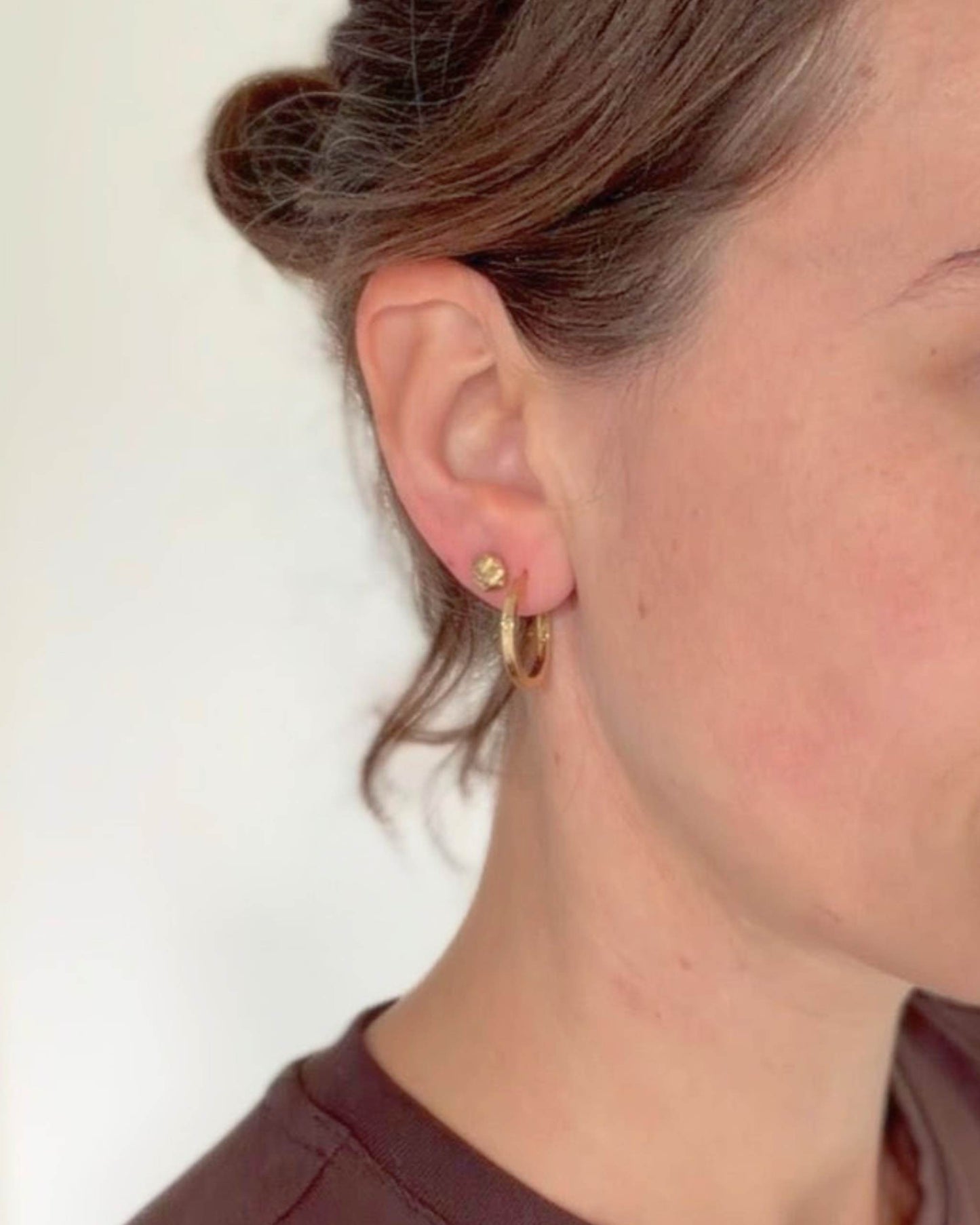 A close-up side profile of a woman wearing a small gold hoop earring and a matching gold stud earring against a plain off-white background.