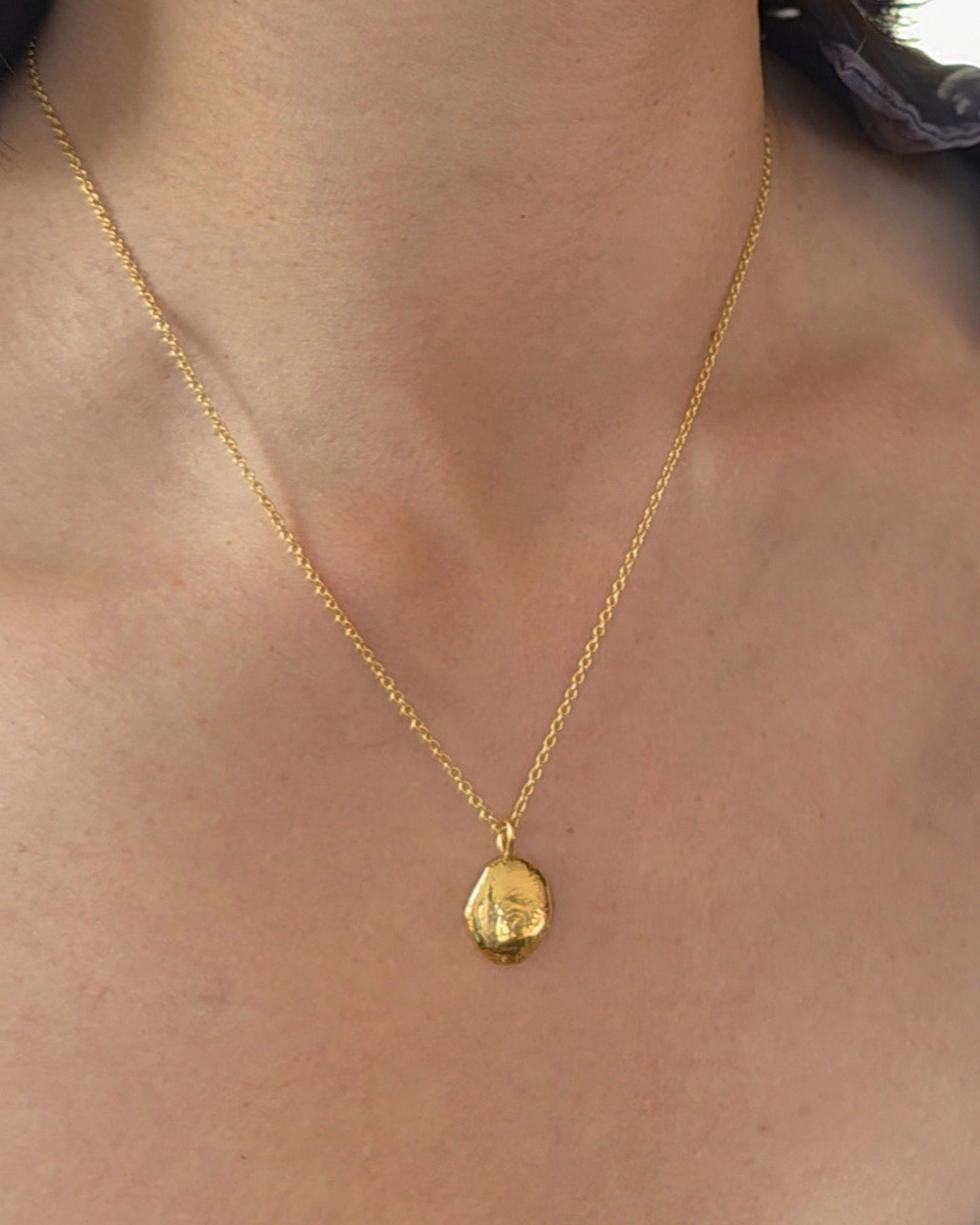A close-up of a person wearing a delicate gold chain necklace with a small, pebble-shaped gold pendant