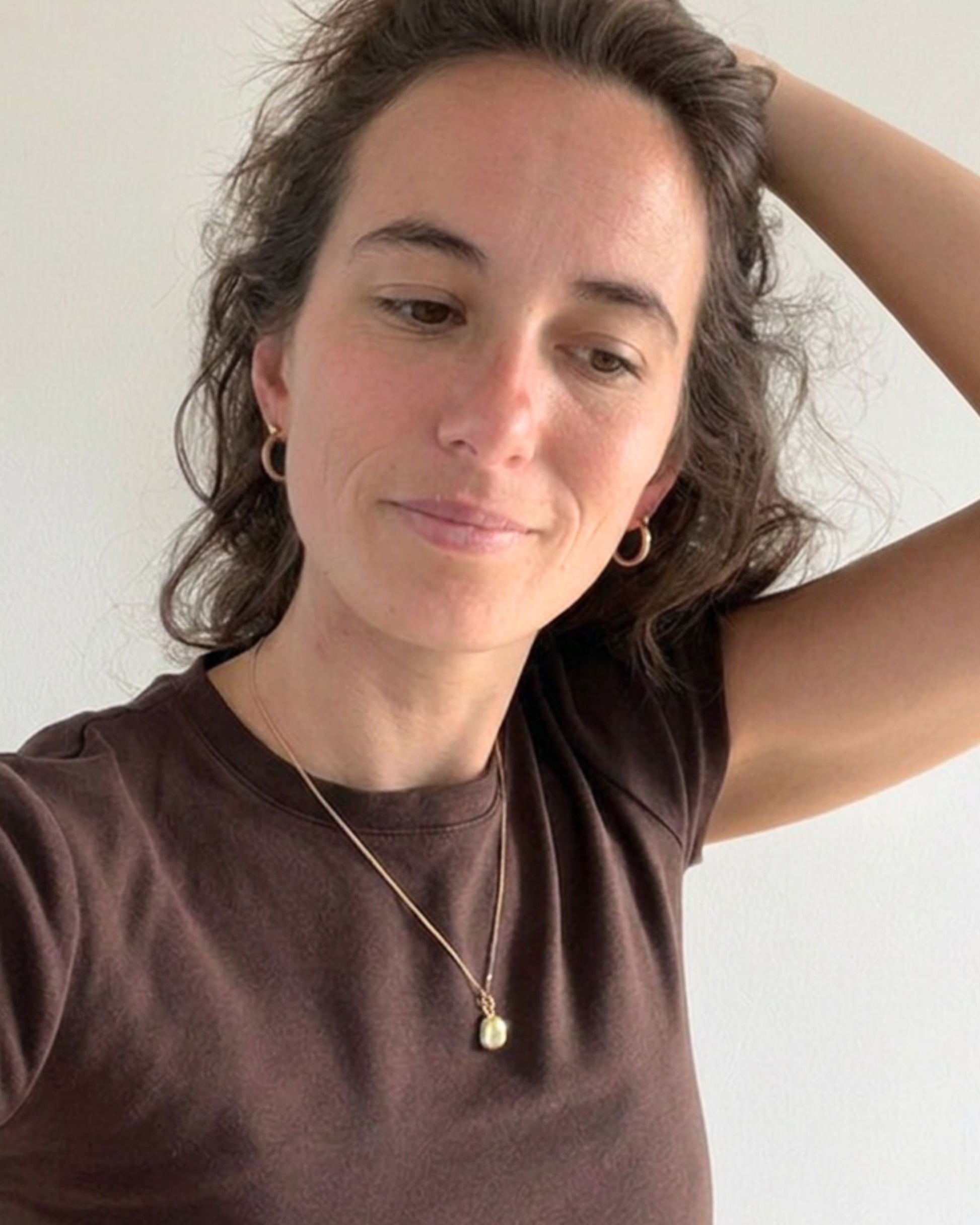 woman with brown hair wearing a dark brown tee-shirt, gold hoop earrings and a fine necklace with a small gold pendant
