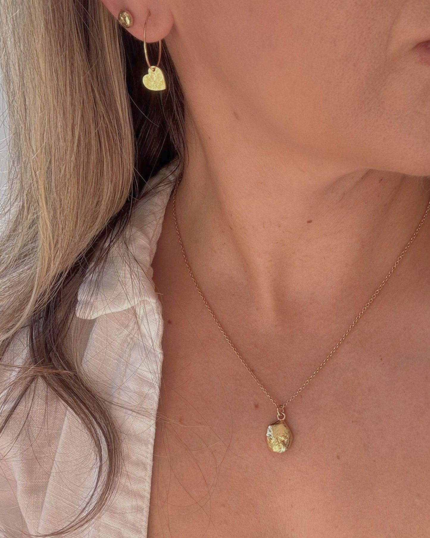 Close-up of a person wearing a gold necklace with an oval pendant and gold heart earrings