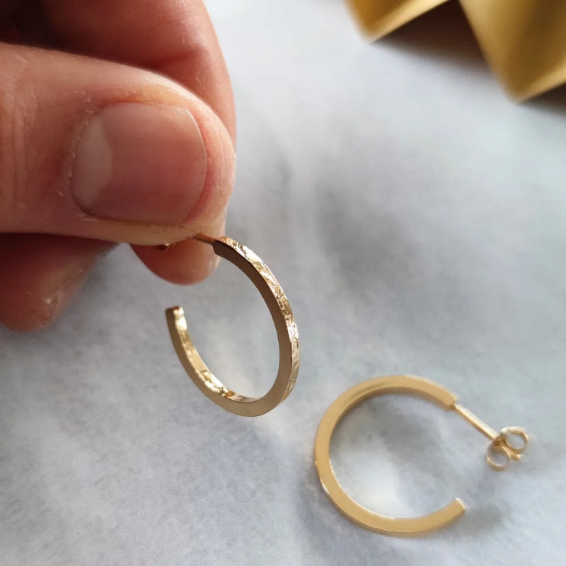 person holding gold texture hoop earrings above marble surface